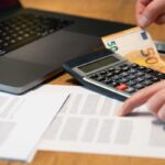 Close-up of hands using calculator with euro notes and laptop on desk.